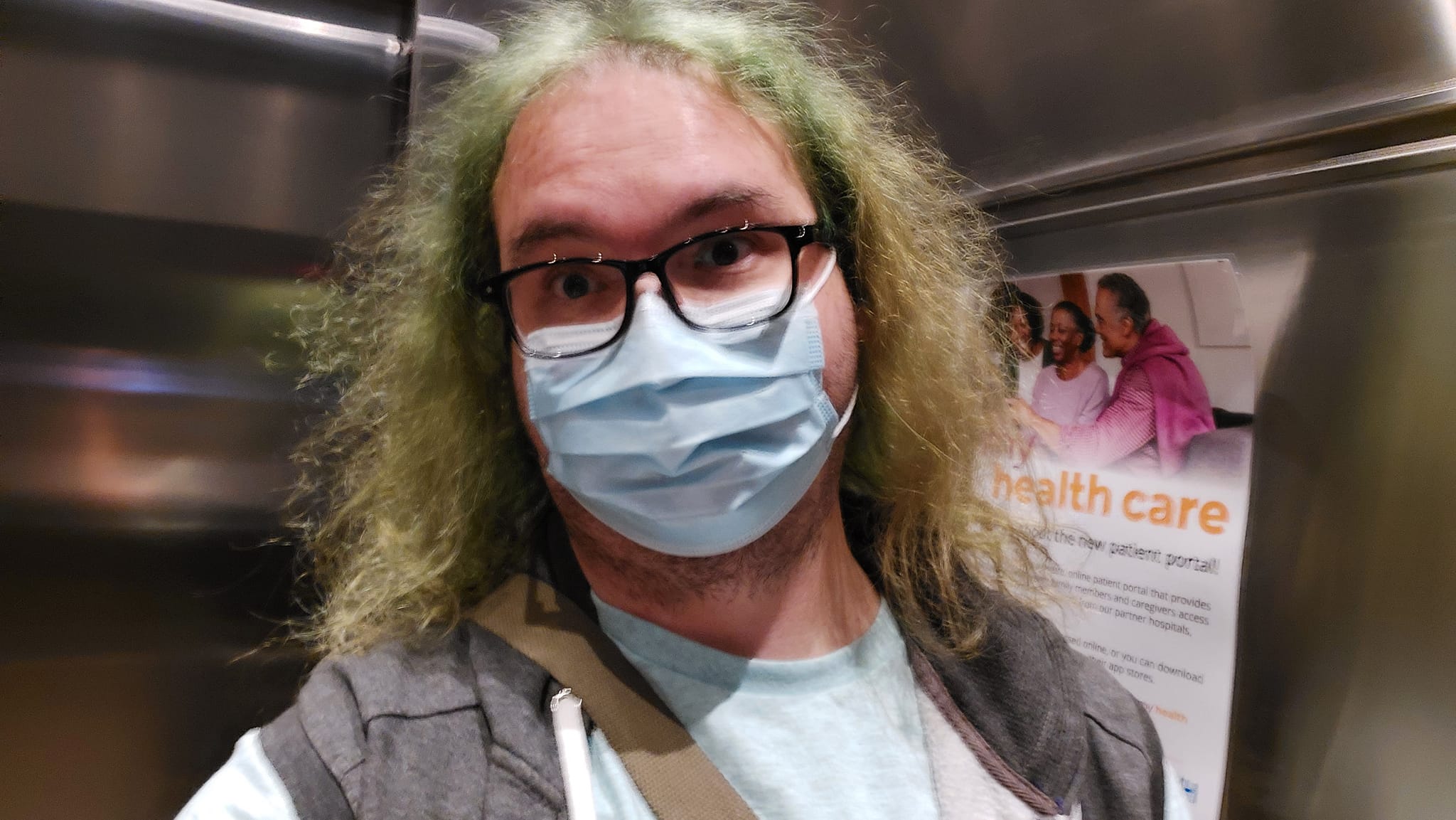 A man with long green hair stands in an elevator with stainless steel walls, he obscures a hospital poster on the wall